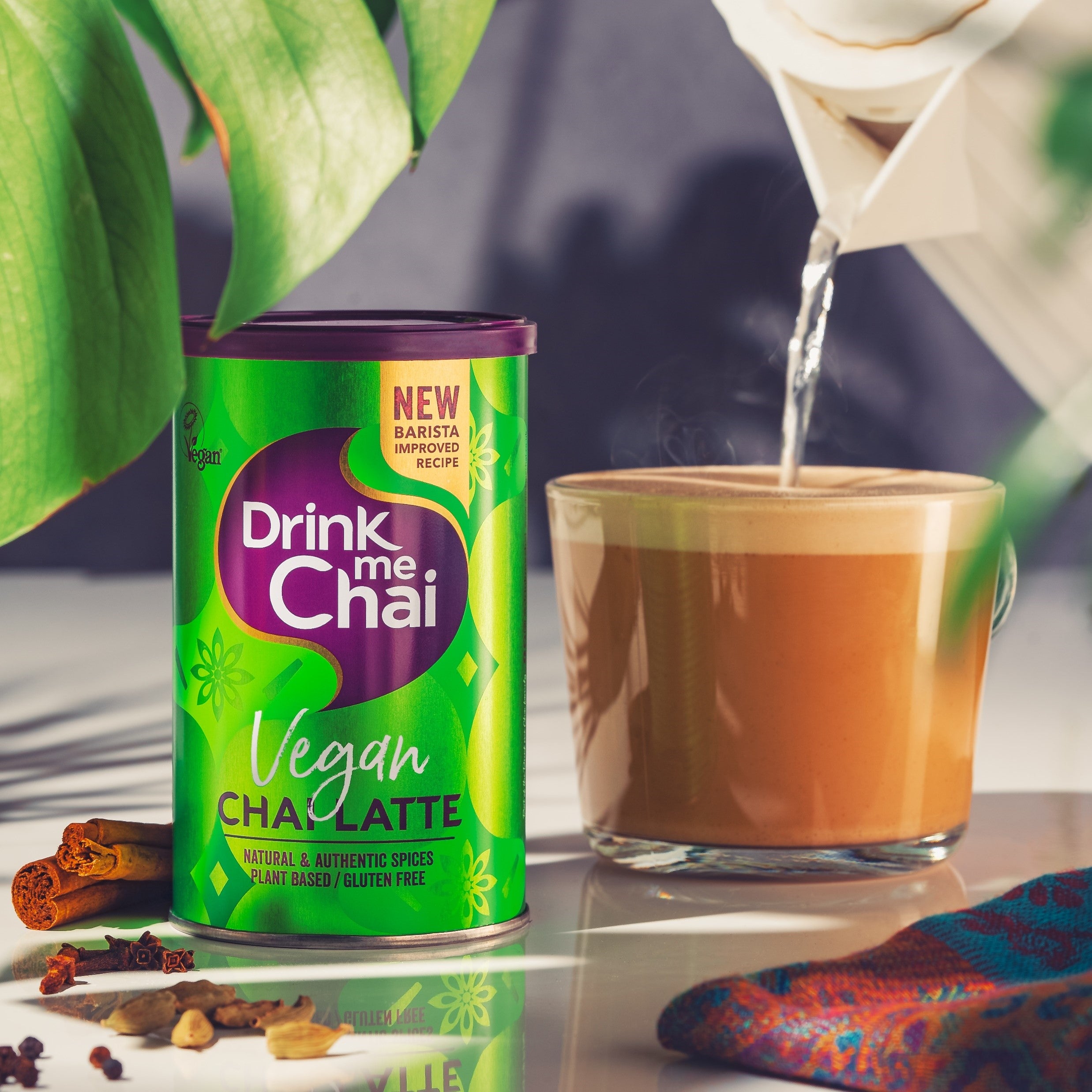 An alternative lifestyle image of a vegan chai latte being poured into a latte glass with a tin of Drink Me Chai in the foreground