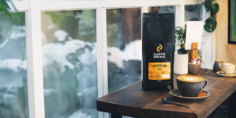 a bag of caffe prima coffee beans stabding on the table next to a steaming cup of coffee