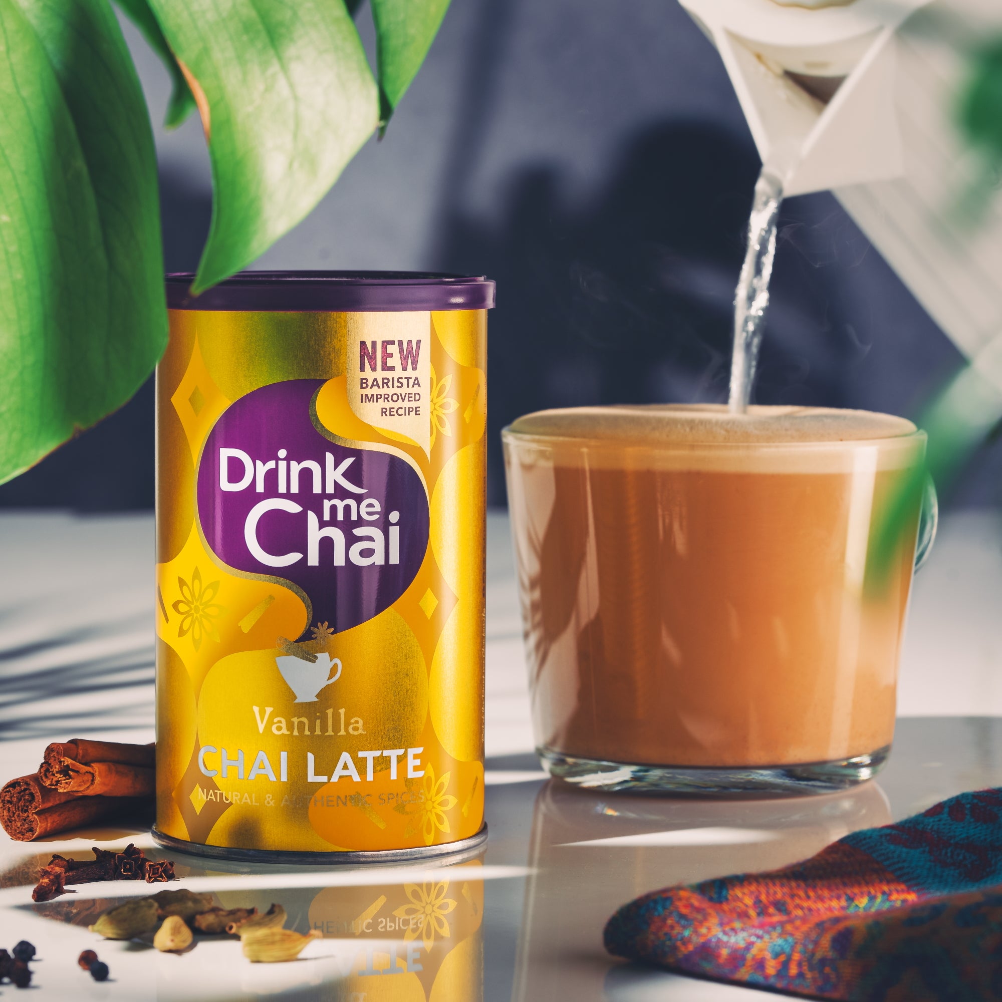 An alternative lifestyle image of a vanilla chai latte being poured into a latte glass with a tin of Drink Me Chai in the foreground